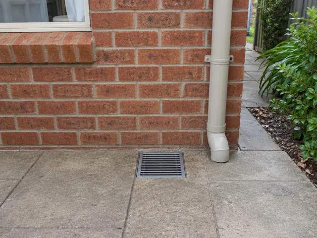 Outdoor drain grate set in dry paved area beside a brick wall and downpipe in Cromer