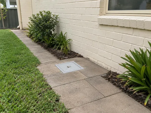 Flush capped access cover set in a paved side path beside a cream brick house in Narrabeen