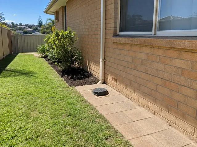 Capped sewer inspection opening on a paved side path beside a brick house and lawn in Dee Why